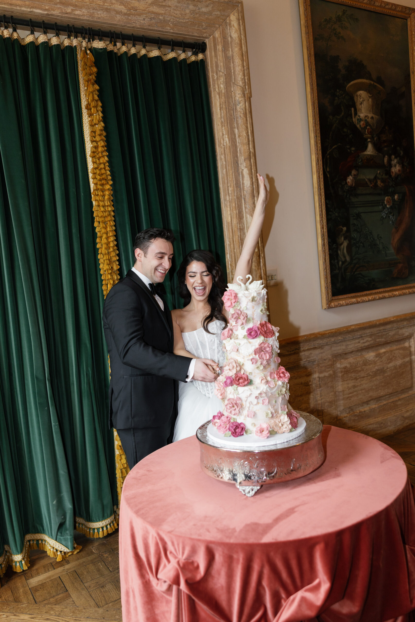 Elegant bride and groom cutting a tall floral wedding cake with pink roses, set against deep green draping and a muted pink cake table at a luxury editorial wedding reception.