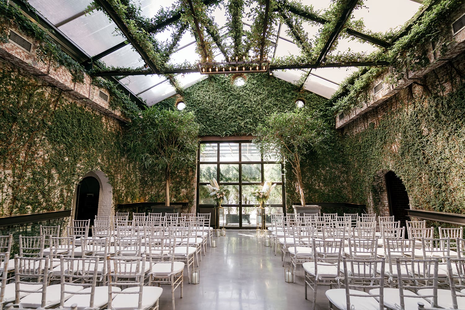 Indoor New York City wedding ceremony venue with ivy-covered brick walls, glass ceiling, white chairs, and modern minimalist aisle setup.
