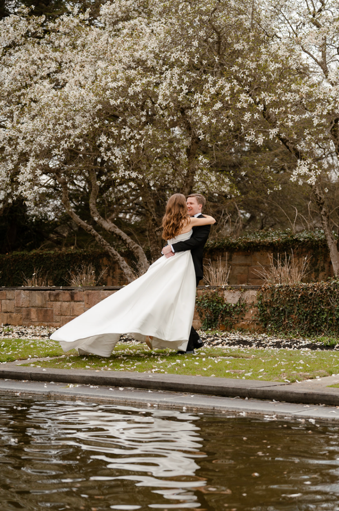 Alt text:
Groom twirling the bride beside a reflecting pool during romantic outdoor wedding portraits at a garden venue.